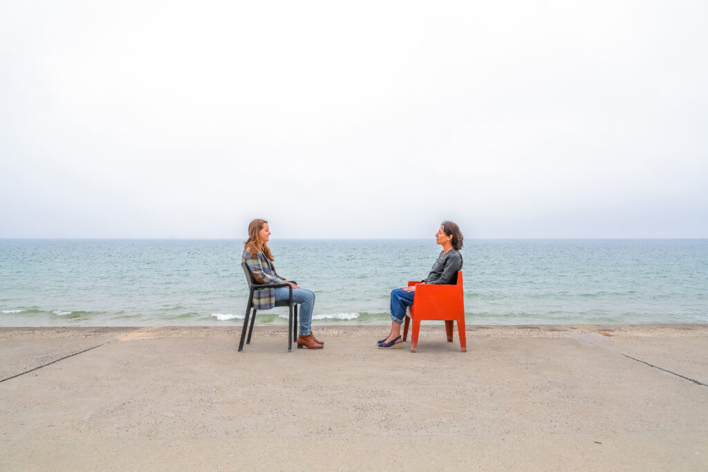 Une photo où deux femmes se tiennent l'une face à l'autre, les yeux clos, mains posées sur les genoux. Elles sont assises sur des chaises, nous les voyons de profil. Elles se trouvent devant la mer, sur la digue de Saint Malo.