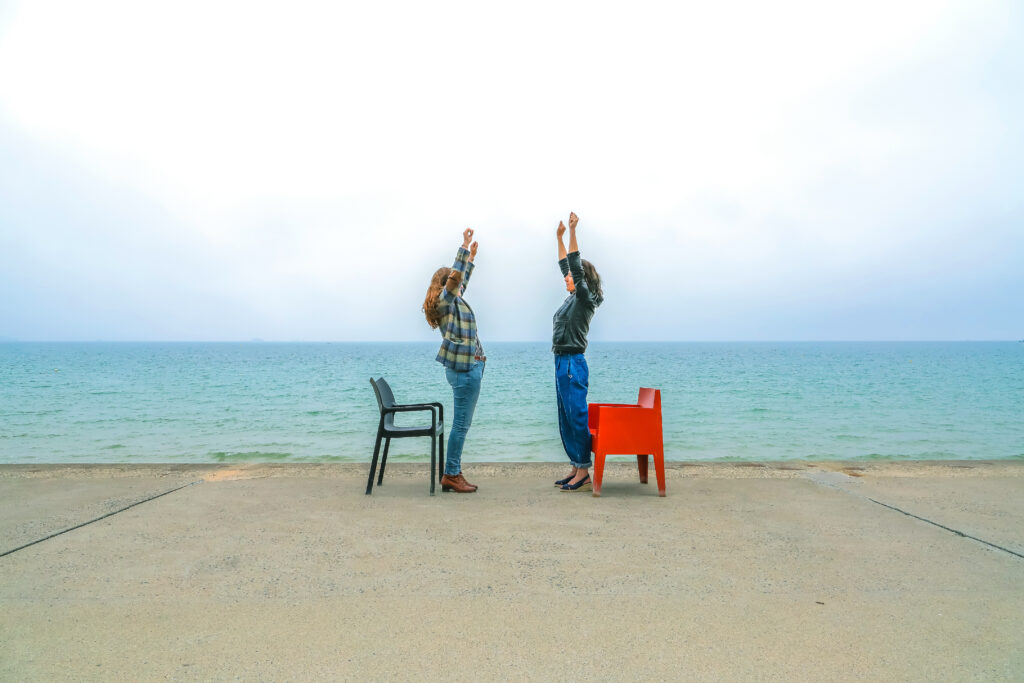 Une photo où deux femmes se tiennent l'une face à l'autre, bras tendus vers le haut. Elles sont debout, de profil, derrière chacune d'elles se trouve une chaise. Elles se trouvent devant la mer, sur la digue de Saint Malo.